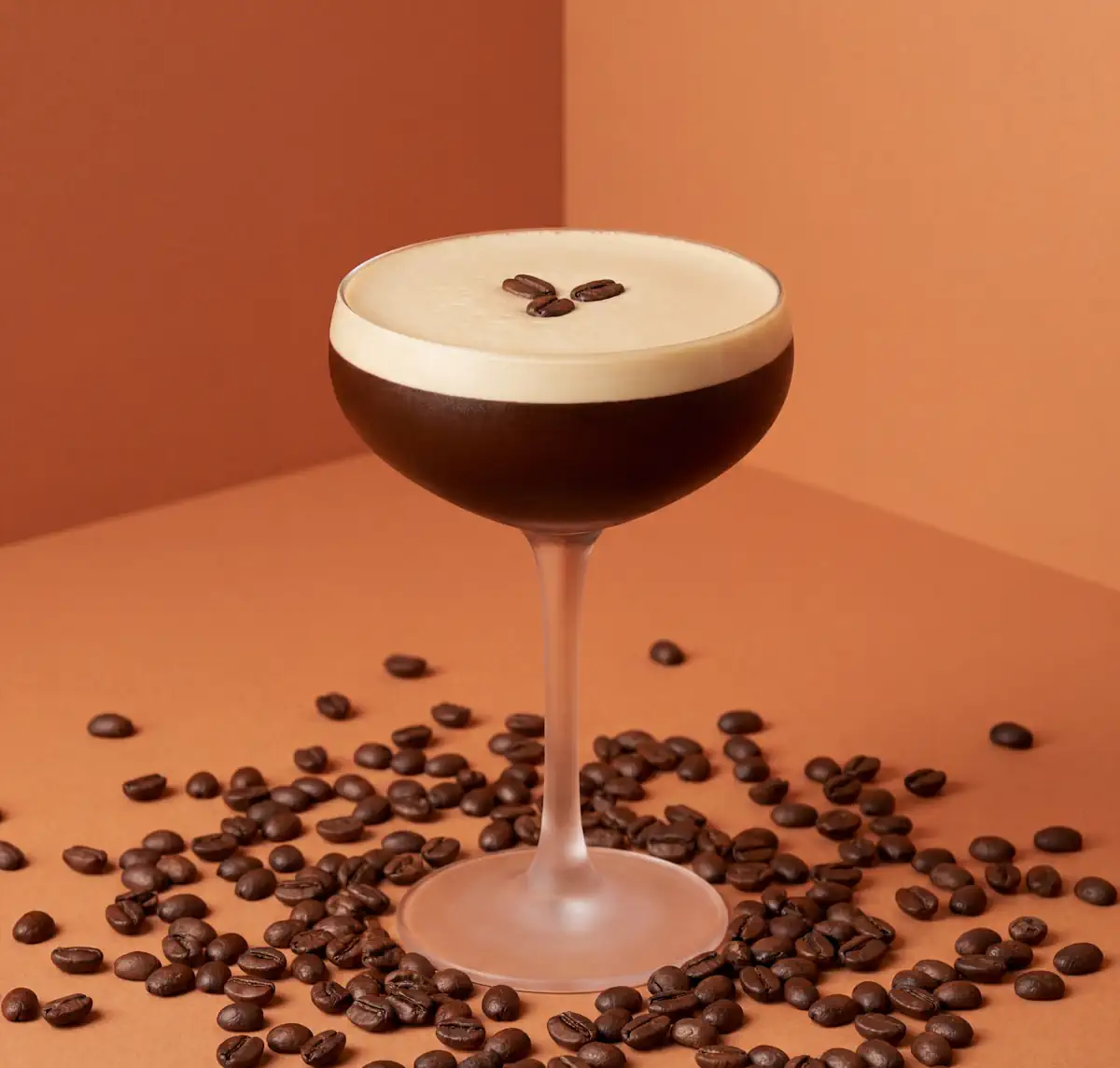 Espresso Martini mit dichter Nitro-Schaumkrone und drei Kaffeebohnen auf einem bernsteinfarbenen Studio-Hintergrund.