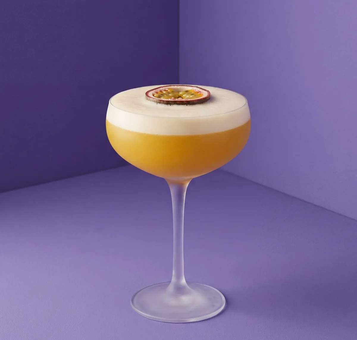 Pornstar Martini Cocktail mit dichter Nitro-Schaumkrone und halber Passionsfrucht als Garnitur vor lila Studio-Hintergrund.